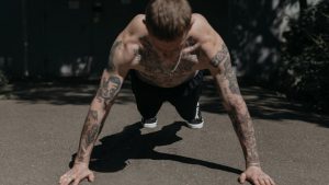 Shirtless tattooed man performing push-ups on a concrete path outdoors, showcasing fitness and endurance.