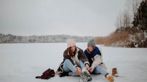 Two women sitting on a frozen lake, putting on ice skates in a winter setting, smiling warmly.