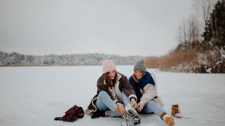 Two women sitting on a frozen lake, putting on ice skates in a winter setting, smiling warmly.